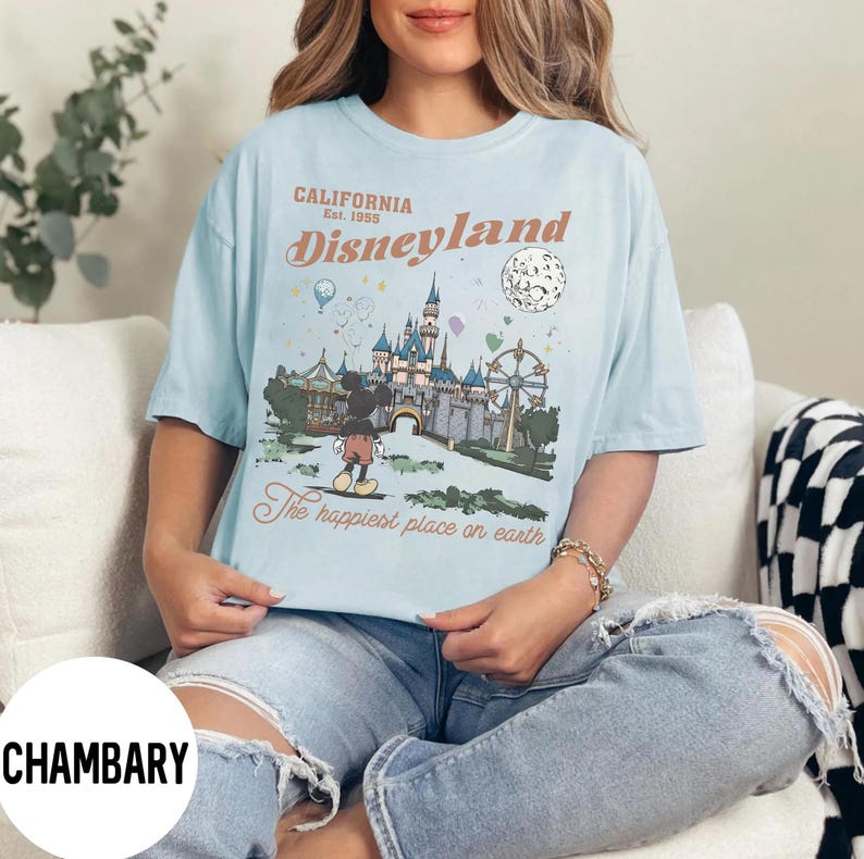 Puede incluir: Camiseta azul claro con un gr&aacute;fico de Disneyland. El dise&ntilde;o incluye el texto "California Est. 1955 Disneyland" y una ilustraci&oacute;n de Mickey Mouse, un castillo y una noria. Tambi&eacute;n se incluye la frase "The happiest place on earth".