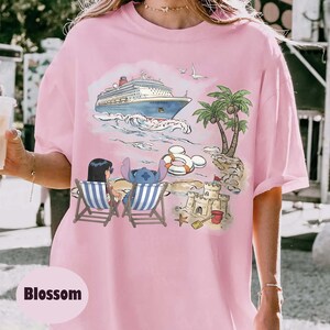 May include: Pink t-shirt featuring a beach scene with a cruise ship, palm trees, and cartoon characters. The design includes a sandcastle, beach chairs, and a life preserver. The word "Blossom" is printed at the bottom.