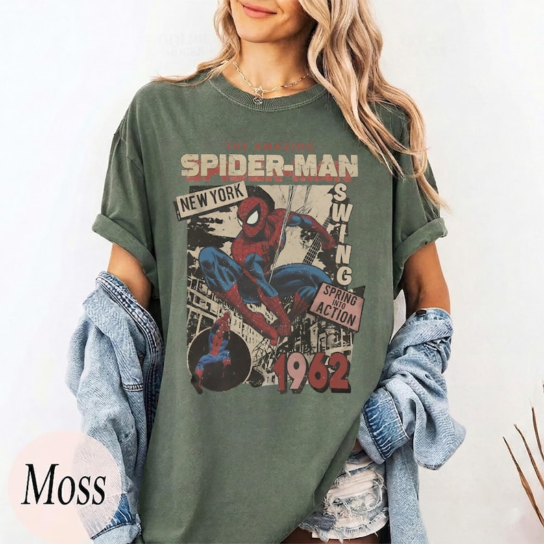 May include: A moss green short-sleeve t-shirt featuring a vintage-style graphic of Spider-Man. The print includes the text THE AMAZING SPIDER-MAN, NEW YORK, SWING, SPRING INTO ACTION, and the year 1962, with a depiction of the character in action.