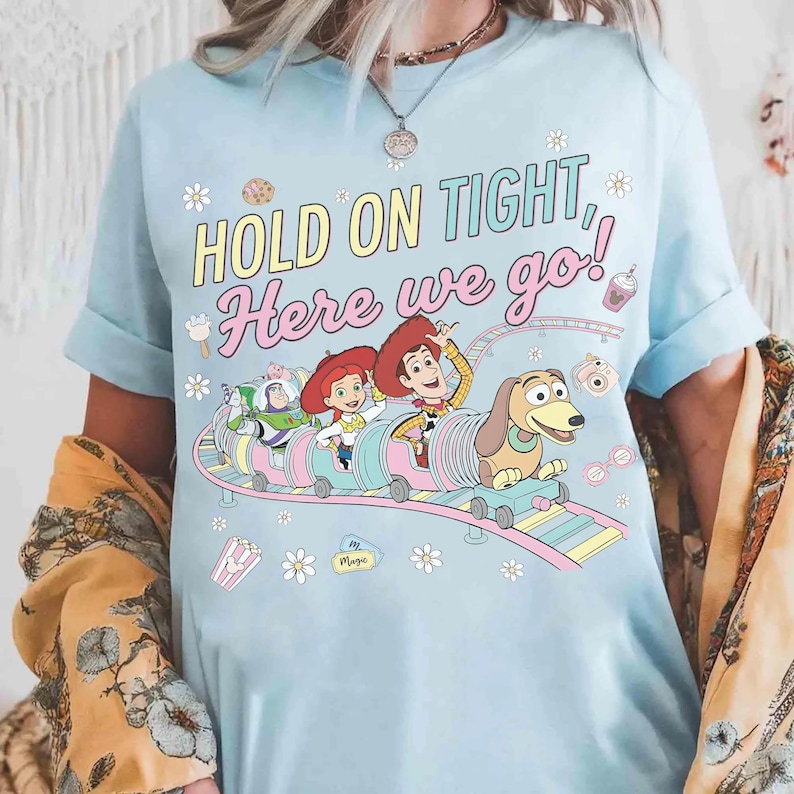 May include: Light blue t-shirt featuring a colorful graphic of characters from the movie Toy Story on a roller coaster. The text "HOLD ON TIGHT, Here we go!" is printed above the graphic. The shirt has rolled-up sleeves.