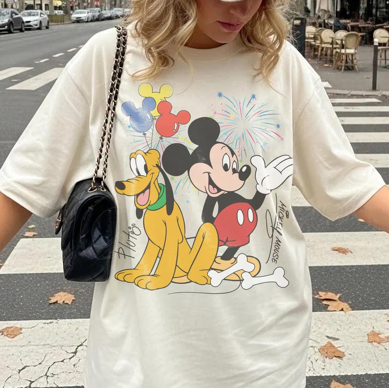 May include: Cream-colored t-shirt featuring Mickey Mouse and Pluto cartoon characters with colorful balloons and fireworks. The words "Mickey Mouse" and "Pluto" are printed on the shirt. The shirt is a casual, oversized fit.