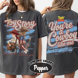 May include: A charcoal gray t-shirt featuring a front graphic of two figures with guitars and the text Toy Story and I knew it, I know you. The back graphic displays the text Toy Story and You're a Cowboy like me in red and white lettering.
