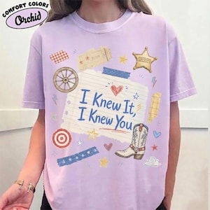 May include: A light purple short-sleeve t-shirt featuring a central graphic of a lined notebook paper with the text I Knew It, I Knew You. The design includes a cowboy boot, a sheriff star, a wagon wheel, a target, and various small decorative icons.
