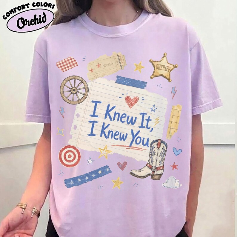 May include: A light purple short-sleeve t-shirt featuring a central graphic of a lined notebook paper with the text I Knew It, I Knew You. The design includes a cowboy boot, a sheriff star, a wagon wheel, a target, and various small decorative icons.