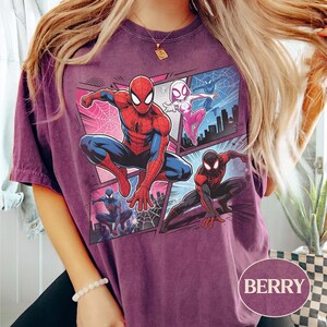 K&ouml;nnte beinhalten: Beerenfarbenes T-Shirt mit einer Comic-Grafik von Spider-Man, Spider-Gwen und Miles Morales. Das Design umfasst Stadtlandschaften und Webmuster. Das T-Shirt hat einen Rundhalsausschnitt und kurze &Auml;rmel. Das Wort "BERRY" ist unten rechts aufgedruckt.