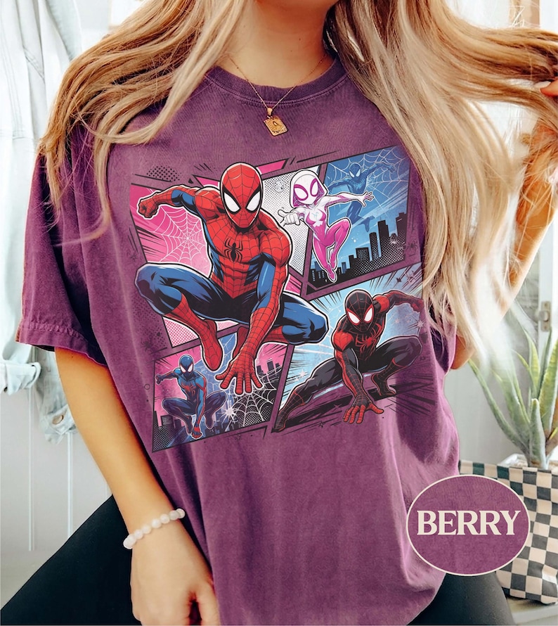 K&ouml;nnte beinhalten: Beerenfarbenes T-Shirt mit einer Comic-Grafik von Spider-Man, Spider-Gwen und Miles Morales. Das Design umfasst Stadtlandschaften und Webmuster. Das T-Shirt hat einen Rundhalsausschnitt und kurze &Auml;rmel. Das Wort "BERRY" ist unten rechts aufgedruckt.