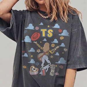 Op de afbeelding: Een antracietgrijs t-shirt met korte mouwen en een grafische print van een persoon met lang haar die gitaar speelt. Het ontwerp bevat blauwe wolken, gele sterren, een rode hoed, een hartvormig koeienprint item en blokken met de letters TS.