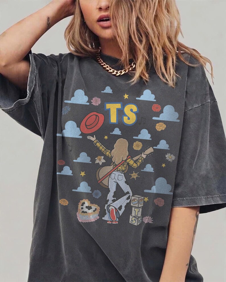 Op de afbeelding: Een antracietgrijs t-shirt met korte mouwen en een grafische print van een persoon met lang haar die gitaar speelt. Het ontwerp bevat blauwe wolken, gele sterren, een rode hoed, een hartvormig koeienprint item en blokken met de letters TS.