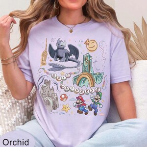 May include: Orchid-colored t-shirt with a colorful graphic design featuring a dragon, castle, Mario and Luigi, and the words "EPIC UNIVERSE". The shirt has a crew neck and short sleeves.