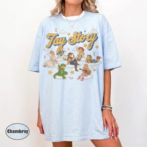 May include: A light blue t-shirt featuring the text "Tay Story" in a yellow cursive font. The graphic includes several illustrated figures dressed in various outfits, including a dinosaur costume, sitting on clouds with musical notes and stars.