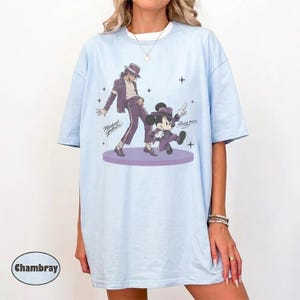 Pode incluir: Uma t-shirt azul-clara com uma estampa gr&aacute;fica de Michael Jackson e Mickey Mouse, ambos vestindo ternos e chap&eacute;us fedora roxos. O design inclui o texto Michael Jackson e Mickey Mouse, com pequenos detalhes de estrelas ao redor das figuras em uma plataforma circular roxa.