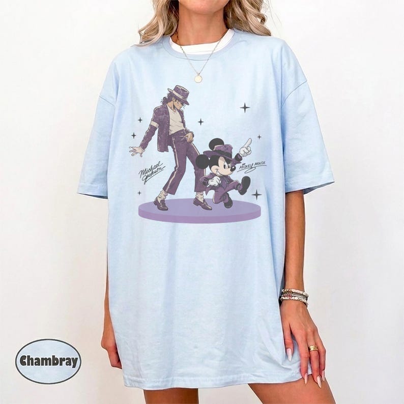Pode incluir: Uma t-shirt azul-clara com uma estampa gr&aacute;fica de Michael Jackson e Mickey Mouse, ambos vestindo ternos e chap&eacute;us fedora roxos. O design inclui o texto Michael Jackson e Mickey Mouse, com pequenos detalhes de estrelas ao redor das figuras em uma plataforma circular roxa.