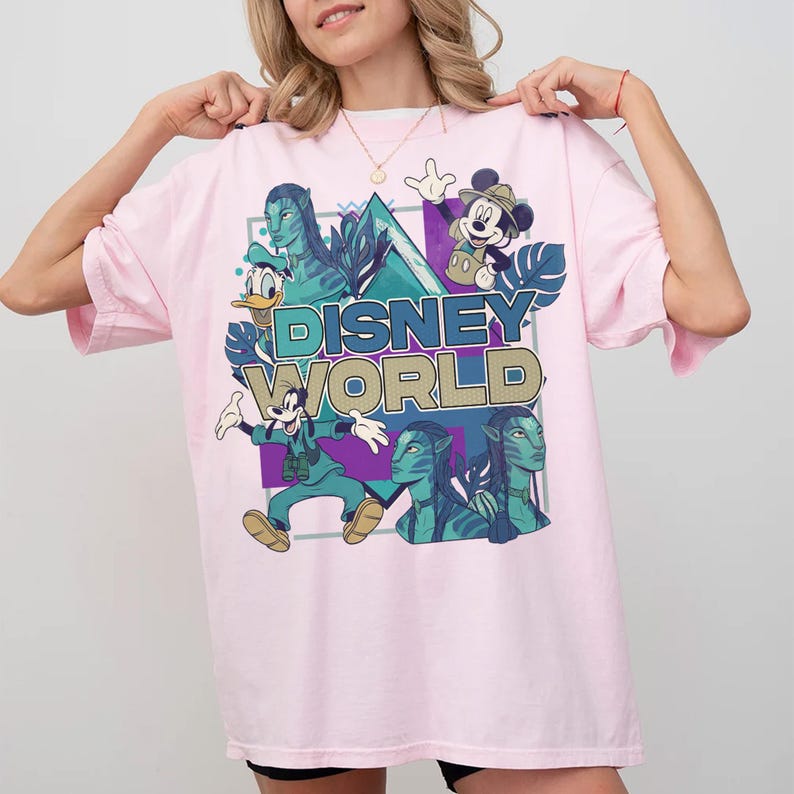 Puede incluir: Una camiseta rosa claro con un estampado gr&aacute;fico que incluye el texto "DISNEY WORLD" en azul y dorado. El dise&ntilde;o presenta ilustraciones de Mickey Mouse, Goofy, Donald Duck y personajes azules de Avatar sobre un fondo geom&eacute;trico.