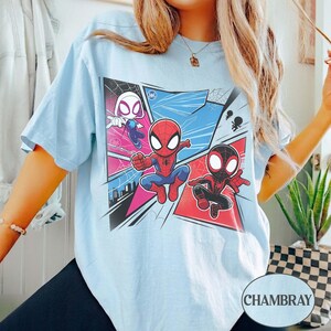 K&ouml;nnte beinhalten: Hellblaues T-Shirt mit einem Comic-Stil-Design von Spider-Man-Charakteren. Die Grafik zeigt Spider-Gwen, Spider-Man und Miles Morales in dynamischen Posen, vor dem Hintergrund von Comic-Panels. Das Wort "CHAMBRAY" ist unten rechts aufgedruckt.