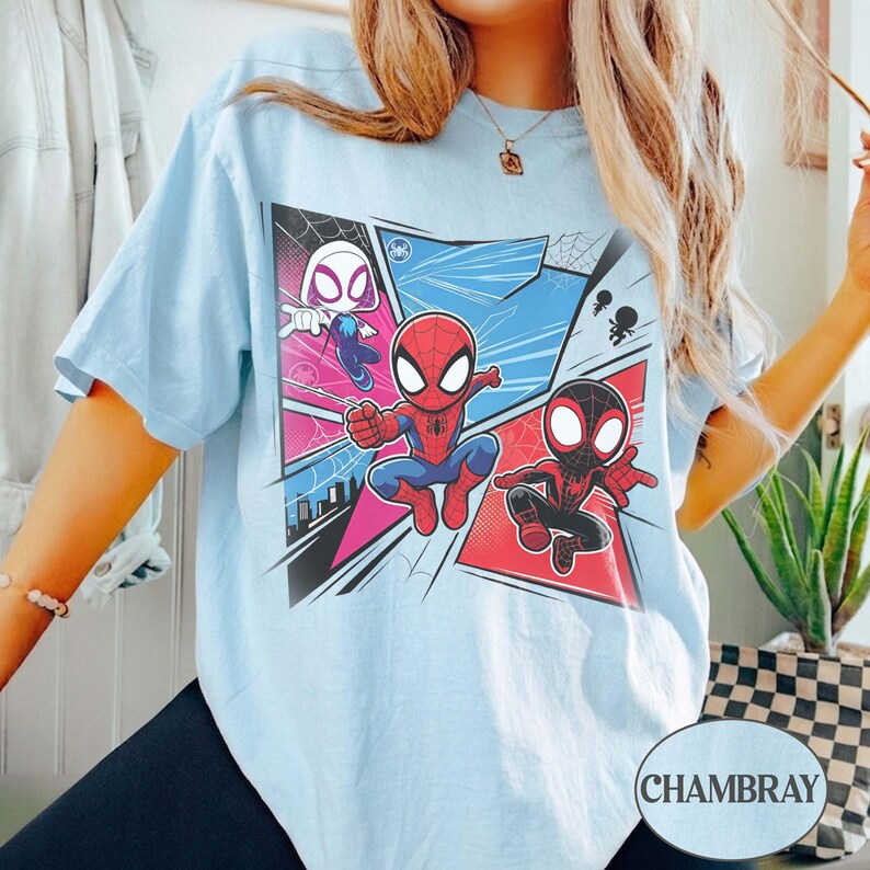 K&ouml;nnte beinhalten: Hellblaues T-Shirt mit einem Comic-Stil-Design von Spider-Man-Charakteren. Die Grafik zeigt Spider-Gwen, Spider-Man und Miles Morales in dynamischen Posen, vor dem Hintergrund von Comic-Panels. Das Wort "CHAMBRAY" ist unten rechts aufgedruckt.