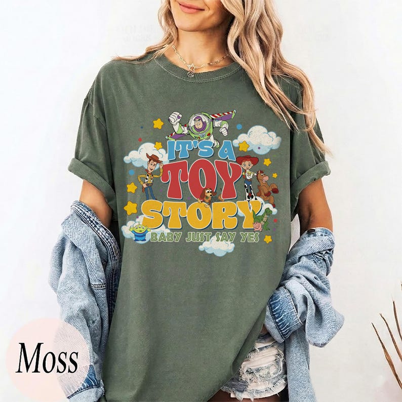 May include: A moss green short-sleeve t-shirt featuring a graphic print of Toy Story characters including Buzz Lightyear, Woody, Jessie, and Bullseye. The text reads IT'S A TOY STORY BABY JUST SAY YES, surrounded by yellow stars and white clouds.