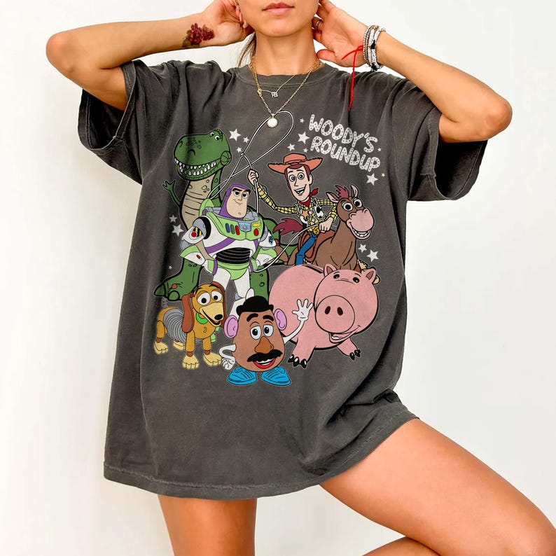 May include: Dark gray t-shirt featuring a colorful graphic of characters from the animated film Toy Story. The design includes Woody, Buzz Lightyear, Rex, Slinky Dog, Mr. Potato Head, Hamm, and the text "Woody's Roundup."