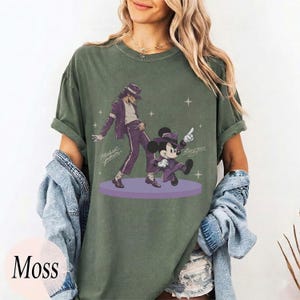 Pode incluir: Uma t-shirt de manga curta verde musgo com uma estampa gr&aacute;fica de Michael Jackson e Mickey Mouse dan&ccedil;ando em ternos roxos num palco circular roxo. O design inclui pequenas estrelas brancas e a assinatura Michael Jackson em letra cursiva.