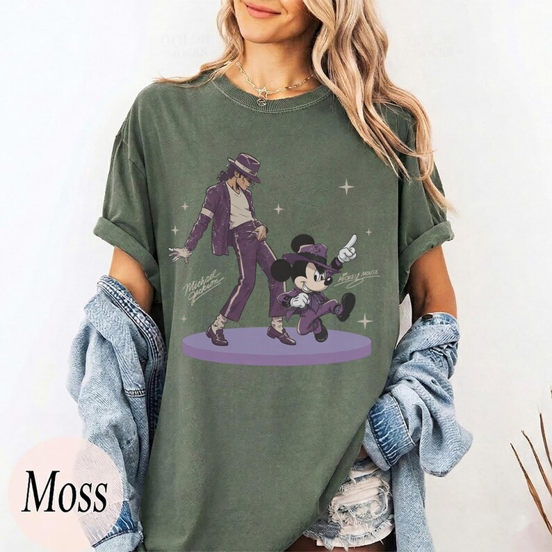 Pode incluir: Uma t-shirt de manga curta verde musgo com uma estampa gr&aacute;fica de Michael Jackson e Mickey Mouse dan&ccedil;ando em ternos roxos num palco circular roxo. O design inclui pequenas estrelas brancas e a assinatura Michael Jackson em letra cursiva.