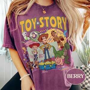 May include: Berry-coloured t-shirt with a Toy Story graphic featuring Woody, Buzz Lightyear, Jessie, and other characters. The design includes the words "TOY STORY" in yellow and green. The shirt is a short-sleeved, casual style.