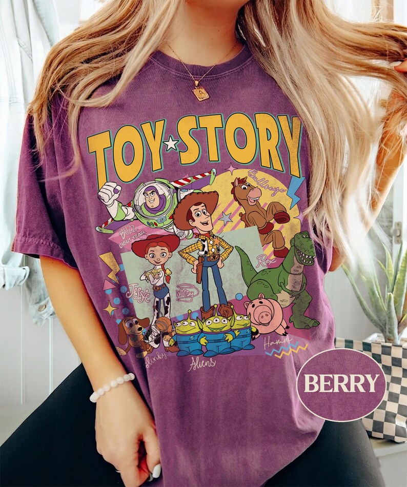 May include: Berry-coloured t-shirt with a Toy Story graphic featuring Woody, Buzz Lightyear, Jessie, and other characters. The design includes the words "TOY STORY" in yellow and green. The shirt is a short-sleeved, casual style.