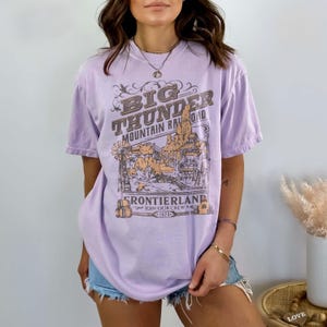 May include: A light purple short-sleeve t-shirt featuring a brown graphic print of a mountain landscape with the text BIG THUNDER MOUNTAIN RAILROAD FRONTIERLAND JOIN OUR CREW 1978. The shirt has a crew neckline and a relaxed fit with short sleeves.