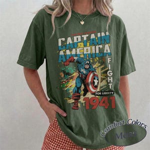 Puede incluir: Una camiseta de manga corta color verde oliva con un gr&aacute;fico de estilo vintage de Capit&aacute;n Am&eacute;rica sosteniendo un escudo. El texto dice CAPTAIN AMERICA, FIGHT FOR LIBERTY y 1941. El dise&ntilde;o incluye colores azul, rojo y blanco en el personaje.