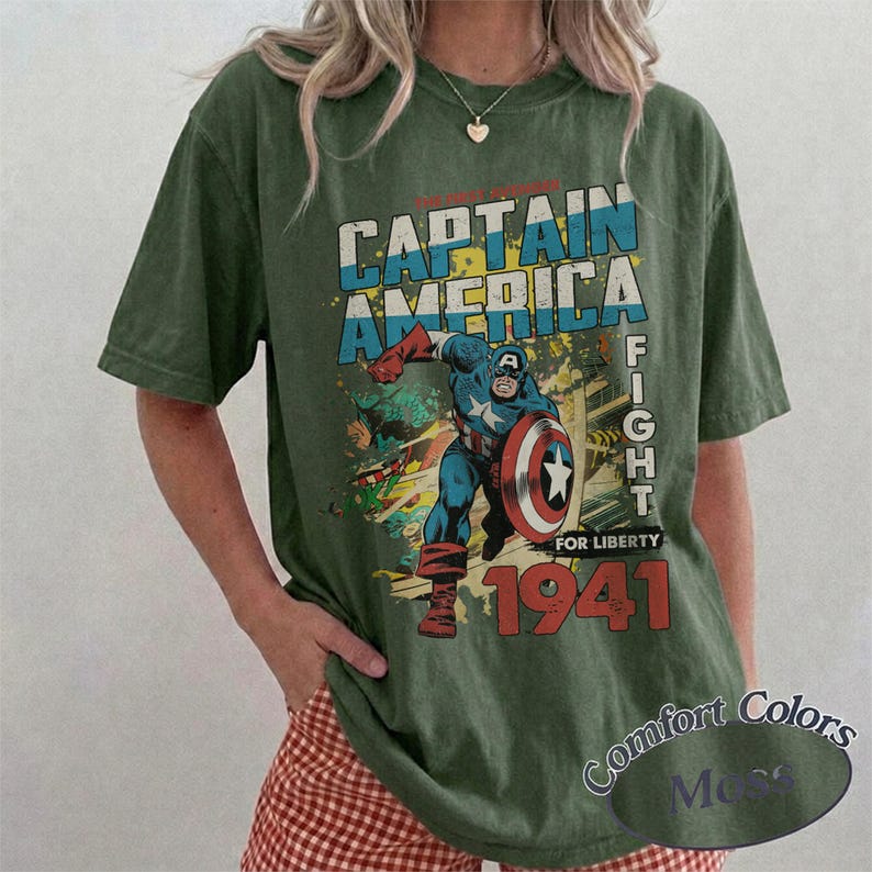 Puede incluir: Una camiseta de manga corta color verde oliva con un gr&aacute;fico de estilo vintage de Capit&aacute;n Am&eacute;rica sosteniendo un escudo. El texto dice CAPTAIN AMERICA, FIGHT FOR LIBERTY y 1941. El dise&ntilde;o incluye colores azul, rojo y blanco en el personaje.