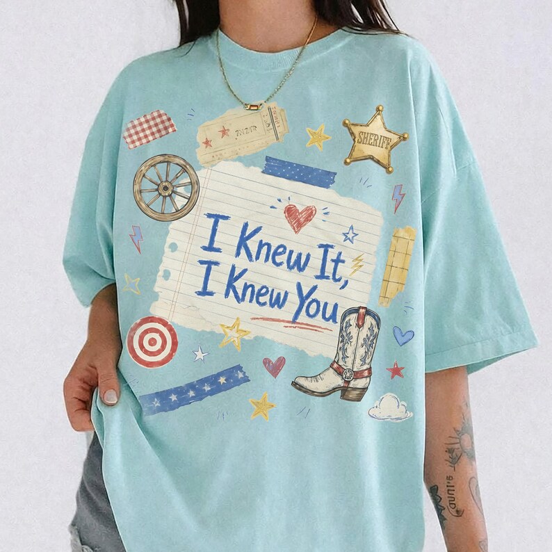 May include: A light blue t-shirt featuring a central graphic of a lined notebook paper with the text "I Knew It, I Knew You" in blue. The design includes a cowboy boot, a sheriff star, a wagon wheel, hearts, stars, and various tape strips.