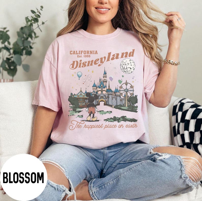 Puede incluir: Camiseta rosa claro con un gr&aacute;fico de Disneyland. El gr&aacute;fico incluye el texto "CALIFORNIA Disneyland Est. 1955" y una ilustraci&oacute;n del castillo, Mickey Mouse y otros elementos del parque. La camiseta tambi&eacute;n tiene el texto "The happiest place on earth."