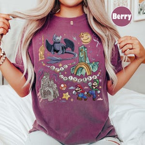 May include: Berry-colored t-shirt with a colorful graphic design. The design includes a dragon, castle, Mario and Luigi, and the words "EPIC UNIVERSE". The shirt has a crew neck and short sleeves.