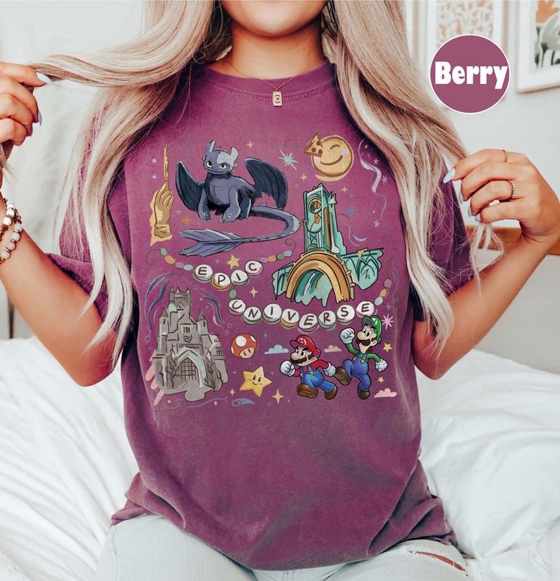 May include: Berry-colored t-shirt with a colorful graphic design. The design includes a dragon, castle, Mario and Luigi, and the words "EPIC UNIVERSE". The shirt has a crew neck and short sleeves.