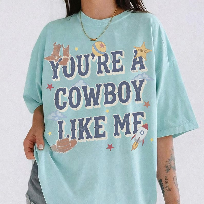 May include: A light blue t-shirt featuring the text "YOU'RE A COWBOY LIKE ME" in dark blue letters. The design includes illustrations of brown cowboy boots, a brown cowboy hat, a yellow sheriff star, a colorful ball, a rocket, and small red stars.