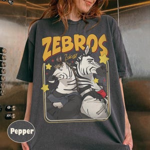 Op de afbeelding: Donkergrijs t-shirt met een cartoon illustratie van twee zebra's in pakken, het woord "ZEBROS" in geel en gele sterren. Het ontwerp is in retro-stijl omlijst. Een casual kledingstuk.