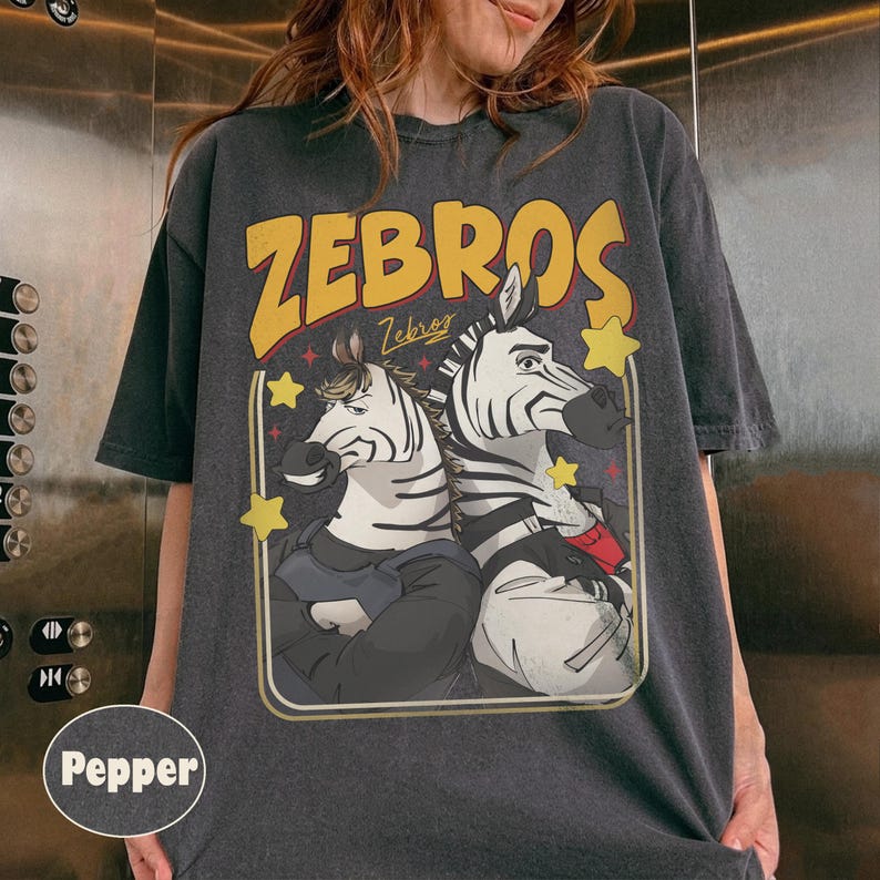 Op de afbeelding: Donkergrijs t-shirt met een cartoon illustratie van twee zebra's in pakken, het woord "ZEBROS" in geel en gele sterren. Het ontwerp is in retro-stijl omlijst. Een casual kledingstuk.