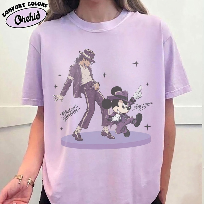 Pode incluir: Uma t-shirt de manga curta em violeta claro com uma impress&atilde;o gr&aacute;fica de Michael Jackson e Mickey Mouse a dan&ccedil;ar numa plataforma circular. Ambas as figuras usam fatos e chap&eacute;us violetas a condizer. O texto Michael Jackson e Mickey Mouse est&aacute; presente.