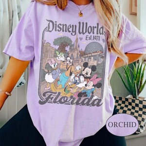 May include: Orchid-colored t-shirt featuring a Disney World graphic with Mickey Mouse, Minnie Mouse, Donald Duck, Daisy Duck, Goofy, and Pluto. The graphic includes the text "Disney World Est. 1971" and "Florida".