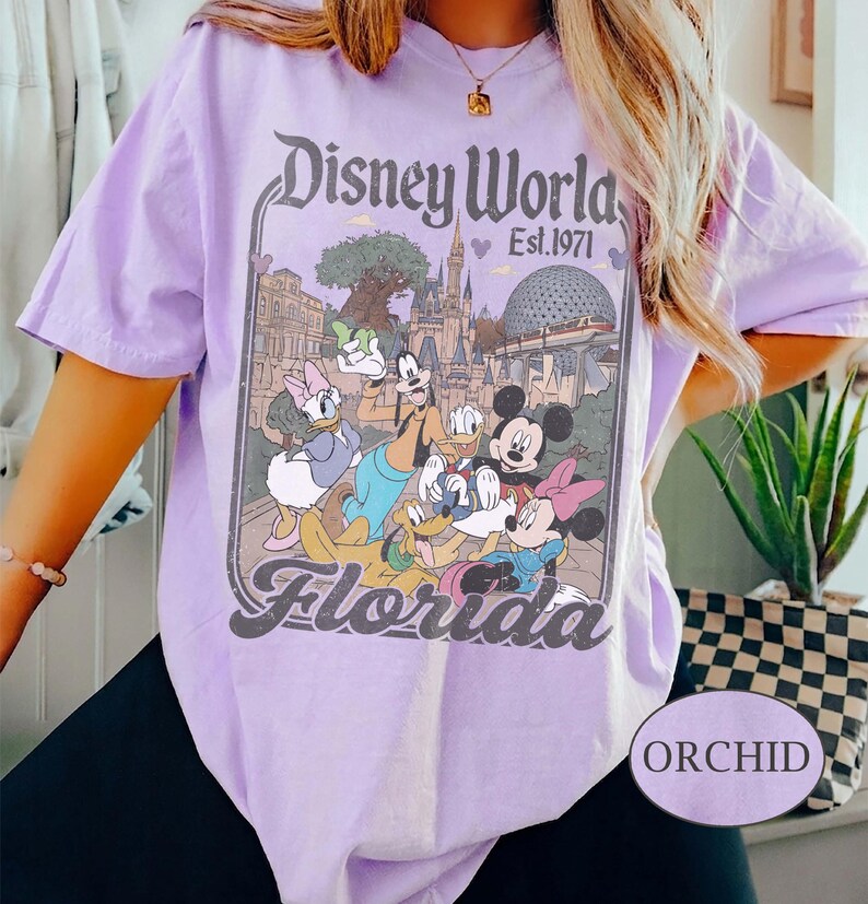 May include: Orchid-colored t-shirt featuring a Disney World graphic with Mickey Mouse, Minnie Mouse, Donald Duck, Daisy Duck, Goofy, and Pluto. The graphic includes the text "Disney World Est. 1971" and "Florida".