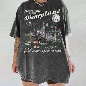 Puede incluir: Camiseta gris oscuro con un gr&aacute;fico de Disneyland. El gr&aacute;fico presenta un castillo, Mickey Mouse y el texto "California Est. 1955 Disneyland The happiest place on earth."