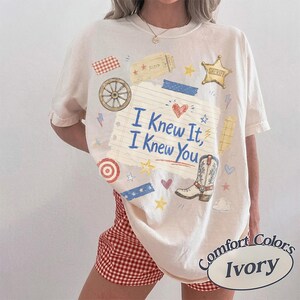 May include: An ivory short-sleeve t-shirt featuring a central graphic of a lined notebook paper with the text "I Knew It, I Knew You" in blue. The design includes a cowboy boot, a sheriff star, a wagon wheel, hearts, and stars in various colors.