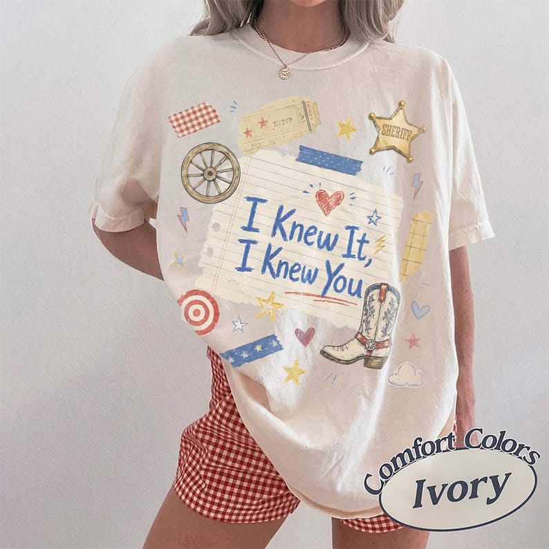 May include: An ivory short-sleeve t-shirt featuring a central graphic of a lined notebook paper with the text "I Knew It, I Knew You" in blue. The design includes a cowboy boot, a sheriff star, a wagon wheel, hearts, and stars in various colors.