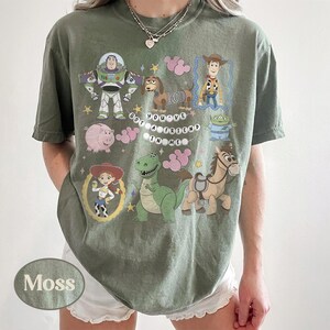 Pu&ograve; includere: T-shirt verde muschio con personaggi dei cartoni animati di Toy Story. Il design include Buzz Lightyear, Woody, Jessie, Rex e altri personaggi. Il testo "YOU'VE GOT A FRIEND IN ME" &egrave; stampato sulla maglietta.