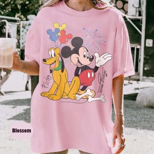 May include: A light pink t-shirt featuring a graphic of Mickey Mouse and Pluto with balloons and fireworks. The text "Mickey Mouse" and "Pluto" are also visible. The shirt is labeled "Blossom".