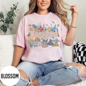 May include: A light pink t-shirt featuring a colorful graphic of Disneyland Resort. The design includes the Disneyland logo and various park attractions. The shirt is worn by a person in blue jeans.