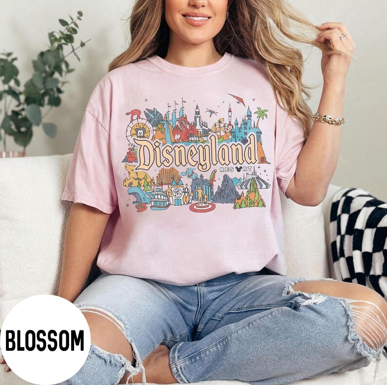 May include: A light pink t-shirt featuring a colorful graphic of Disneyland Resort. The design includes the Disneyland logo and various park attractions. The shirt is worn by a person in blue jeans.