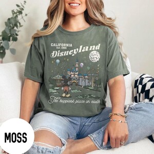 Puede incluir: Camiseta verde musgo con un gr&aacute;fico de Disneyland. El dise&ntilde;o incluye el texto "CALIFORNIA Est. 1955 Disneyland" y una ilustraci&oacute;n del castillo, Mickey Mouse y otros elementos del parque. Tambi&eacute;n incluye la frase "The happiest place on earth".