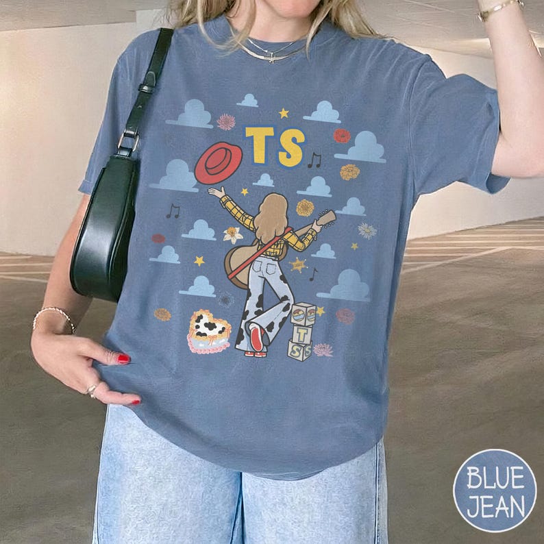 Op de afbeelding: Een blauw t-shirt met een afbeelding van een persoon met lang haar in een geel geruit overhemd en een broek met koeienprint, die een gitaar vasthoudt. Het ontwerp bevat een rode hoed, de letters TS, wolken, sterren, muzieknoten en blokken met TS.