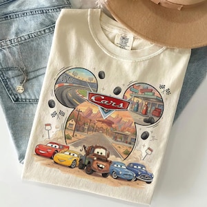 Puede incluir: Una camiseta de color crema con un estampado gr&aacute;fico del logotipo de Disney Pixar Cars y personajes como Rayo McQueen, Mate y otros veh&iacute;culos. El dise&ntilde;o incluye un circuito de carreras, un pueblo del desierto y banderas a cuadros.