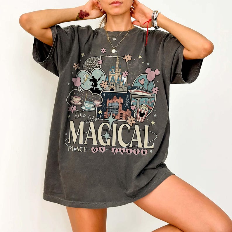 Puede incluir: Camiseta gris oscuro con un gr&aacute;fico colorido con el texto "The Most Magical Place on Earth". El dise&ntilde;o incluye ilustraciones de un castillo, tazas de t&eacute;, un monorra&iacute;l y palomitas de ma&iacute;z.