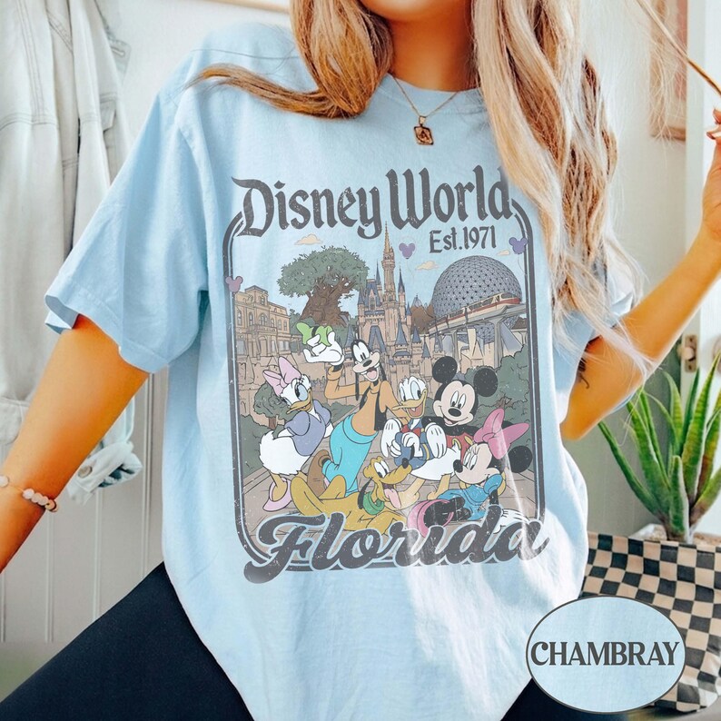 May include: Light blue t-shirt featuring a vintage-style Disney World graphic with Mickey Mouse, Donald Duck, and other characters. The graphic includes the text "Disney World Florida Est. 1971". The shirt is a chambray color.